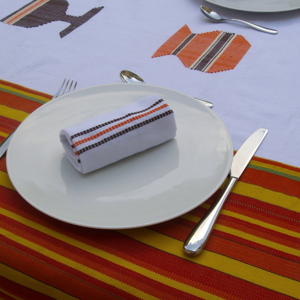 nappe avec motif ethnique, art naif une coupe de vin et une cruche. la nappe est blanche avec des bandes dans les tons ornagés sur les cotés et au centre les motifs en broderies. Camaieux orange, jaune et marron. Serviettes assorties
